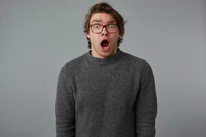 Portrait of young amazed guy with glasses, stands over gray background with wide open mouth and eyes, with surprised expression, looks scared and shocked. photo
