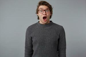 Photo of young shocked man with glasses, stands over gray background with wide open mouth and eyes, with surprised expression, looks scared and shocked.
