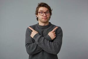 Photo of young confused pensive man with glasses wears in gray sweater, stands over gray background, shows fingers in different directions.