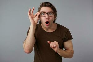 Young wondered with glasses wears in basic t-shirt, stands over gray background and broadly smiles, looks looks trough glasses with wide open mouth and eyes. photo