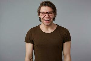 foto de joven riendo hermoso hombre con lentes usa en básico camiseta, soportes terminado gris antecedentes y en general sonrisas, mira contento y se preguntó.