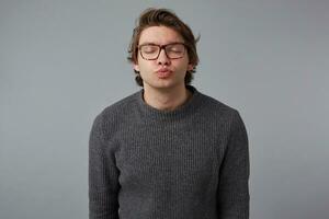 Photo of young guy with glasses wears in gray sweater, stands over gray background and sends kiss to the camera with closed eyes.