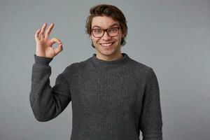 Photo of young cheerful handsome guy with glasses wears in gray sweater, stands over gray background and broadly smiles, looks happy and glad, shows okey gesture.