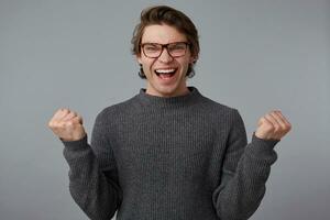 retrato de joven alegre hombre con lentes usa en gris suéter, soportes terminado gris antecedentes. en general sonrisas y aprieta puños, won el coche y siente felicidad. foto