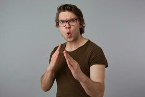 Photo of young guy with glasses wears in blank t-shirt standing in defensive posture, ready to punch, stands over gray background and try look expressing anger and fury.