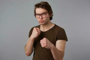 Photo of aggressive young man guy with glasses wears in blank t-shirt standing in defensive posture, keeping fists clenched, ready to punch, stands over gray background.