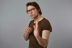 Young severe man with glasses wears in blank t-shirt standing in defensive posture, ready to punch, stands over gray background and looks expressing anger and fury. photo