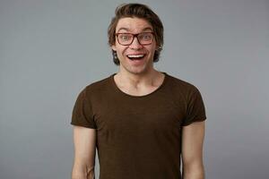 Photo of young happy amazed handsome man with glasses wears in basic t-shirts, stands over gray background and broadly smiles, looks happy and glad.