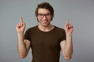 Portrait of young happy man with glasses, stands over gray background with surprised expression, points fingers up at a copy space over his head, looks at the camera and broadly smiles. photo