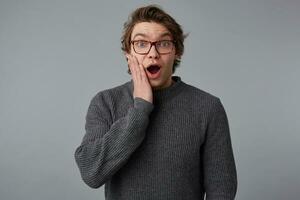 Photo of young shocked guy with glasses wears in gray sweater, stands over gray background and touches cheek, with wide open mouth and eyes, with surprised expression.