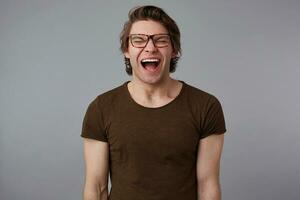 Photo of young crying guy with glasses wears in blank t-shirt, stands over gray background and looks unhappy and sad.