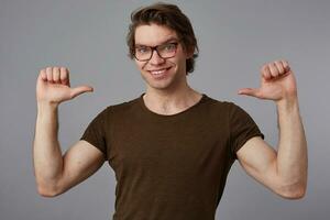 Young cheerful guy with glasses wears in blank t-shirt standing over gray background and pointing at himself, looks happy and broadly smiles and says I'm cool. photo