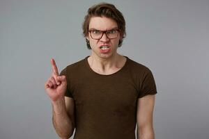 Photo of young angry guy with glasses wears in blank t-shirt, stands over gray background and looks at the camera with dissatisfied expression.