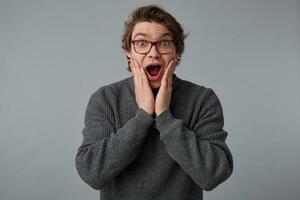 Wow, unbelievable news Young shocked guy with glasses, stands over gray background with wide open mouth and eyes, touches cheeks, with surprised expression. photo