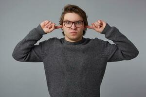 Portrait of young angry man with glasses wears in gray sweater, stands over gray background, doesn't want to listen, disturbed by noise and covers ears with fingers. photo