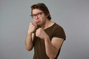 Young guy with glasses wears in blank t-shirt standing in defensive posture, keeping fists clenched, ready to punch, stands over gray background and try look expressing anger and fury. photo
