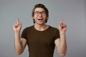 Portrait of young happy amazed man with glasses, stands over gray background with surprised expression, points fingers up at a copy space over his head. photo