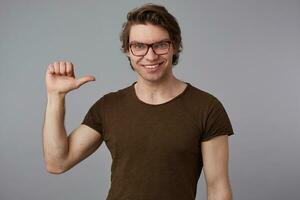 Young cool man with glasses wears in blank t-shirt standing over gray background and pointing at himself, looks cheerful and broadly smiles. photo