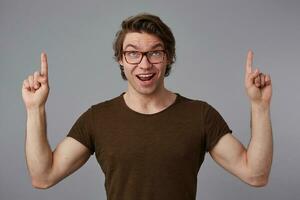 Portrait of young happy amazed guy with glasses, stands over gray background with surprised expression, points fingers up at a copy space over his head, looks at the camera and broadly smiles. photo