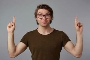 Young happy guy with glasses, stands over gray background with surprised expression, points fingers up at a copy space over his head, looks up and smiles. photo