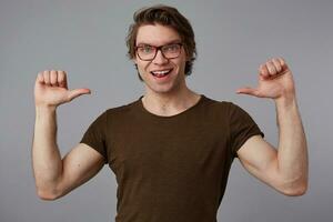 Positive man with glasses wears in blank t-shirt standing over gray background and pointing with fingers at himself, looks cheerful and broadly smiles and says I'm cool. photo