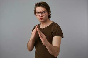 Young man with glasses wears in blank t-shirt standing in defensive posture, ready to punch, stands over gray background and looks expressing anger and fury. photo