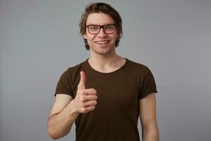 Portrait of young happy man with glasses, stands over gray background with cheerful expression, shows like gesture and broadly smiles. photo