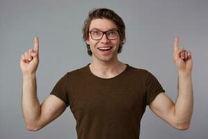 Portrait of young cheerful guy with glasses, stands over gray background with surprised expression, points fingers up at a copy space over his head, looks at the camera and broadly smiles. photo