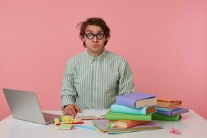 Young guy with glasses, wears on blank shirt, sitting at a table with books, working at a laptop, looks shocked and confused. Isolated over pink background. photo