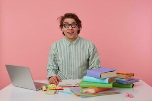 Young happy amazed man with glasses, wears on blank shirt, sitting at a table with books, working at a laptop, looks surprised, hears cool news. Isolated over pink background. photo