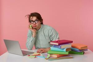 Young sad guy with glasses, wears on blank shirt, sitting at a table with books, working at a laptop, looks unhappy and bored, looks at the camera isolated over pink background. photo