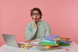 Studio shot of young wondered guy with glasses, wears on blank shirt, sitting at a table with books, working at a laptop, looks surprised and shocked. Isolated over pink background. photo