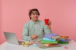 young happy man with glasses, sitting at a table with books, working at a laptop, looks glad, wears on blank shirt, looks at the camera and holds cup of coffe isolated over pink background. photo
