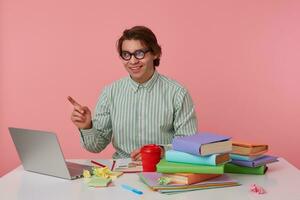 Portrait of young smiling man with glasses, sitting at a table with books, working at a laptop, looks cheerful, looks and pointing with fingers to the left side, isolated over pink background. photo
