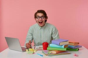 young cheerful man with glasses, sitting at a table with books, working at a laptop, looks glad, wears on blank shirt, looks at the camera with happy expression, isolated over pink background. photo