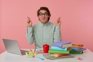 retrato de hombre con anteojos, sentado a un mesa con libros, trabajando a un computadora portátil, mira arriba con cruzado dedos, esperanzas para bueno suerte, aislado terminado rosado antecedentes. foto