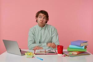 Photo of young thinking guy with glasses, sitting at a table with books, working at a laptop, wishful looks up, isolated over pink background.