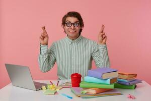 Photo of young guy with glasses, sitting at a table with books, working at a laptop, looks at the camera and smiling, with crossed fingers, hopes for good luck, isolated over pink background.