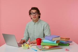 Portrait of man with glasses, sitting at a table with books, working at a laptop, looks at the camera with disgusted expression, isolated over pink background. photo