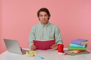 Photo of young serious guy with glasses, sitting at a table with books, working at a laptop, concentrate looks at the camera, with opened books in heands, isolated over pink background.