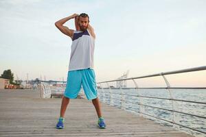 Young sporty bearded guy doing a warm-up after a morning run at the seaside, leads healthy active lifestyle, looks away, feals great. photo