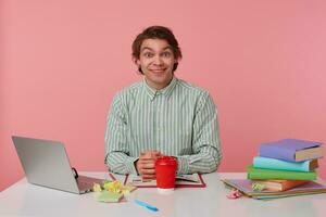 Photo of young cheerful guy with glasses, sitting at a table with books, working at a laptop, looks at the camera and smiling, isolated over pink background.