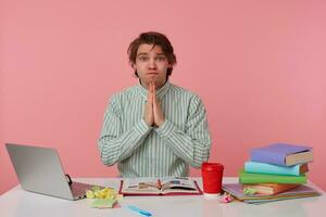 Photo of young guy with glasses, sitting at a table with books, working at a laptop, looking imploringly at the camera with prayer gesture, isolated over pink background.