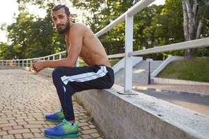 Young attractive bearded man has extreme sport in park, rest at the park after jogging, leads healthy active lifestyle, looks at the camera. Fitness male model. photo