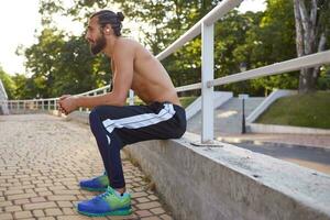 joven cansado deportivo barbado hombre tiene extremo deporte en parque, descanso después correr, Guías sano activo estilo de vida, mira lejos. aptitud masculino modelo. foto