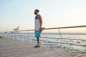 Photo of young sporty bearded man at the seaside doing a warm-up before a morning run, leads healthy active lifestyle, looks away. Fitness male model.