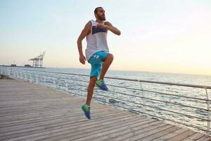 Young sporty handsome bearded jumping guy doing morning exercises by the sea, warm-up before run, leads healthy active lifestyle. Fitness and sport concept. photo