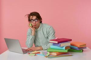 Young bored guy with glasses,sitting at a table with books, working at a laptop, looks unhappy, sadly looks up and dreams of leaving home as soon as possible, isolated over pink background. photo