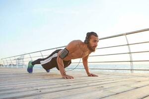 Young sporty bearded guy doing morning exercises by the sea, warm-up after run, doing pushups, keeps the plank, listening favorite songs on headphones, leads healthy active lifestyle. photo