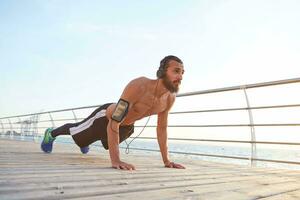 Young sporty bearded guy listening favorite songs on headphones, doing morning exercises by the sea, doing pushups, keeps the plank, warm-up after run, leads healthy active lifestyle. photo